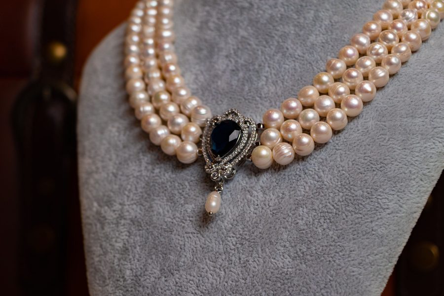 A triple-strand pearl necklace with a sapphire and diamond clasp, shown on a grey bust.