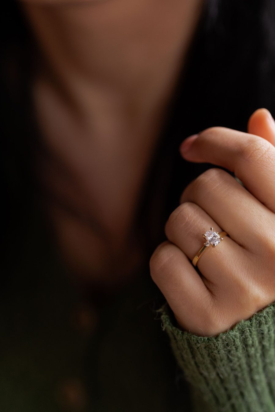 A hand wearing a slim gold solitaire ring, resting against a soft dark background.