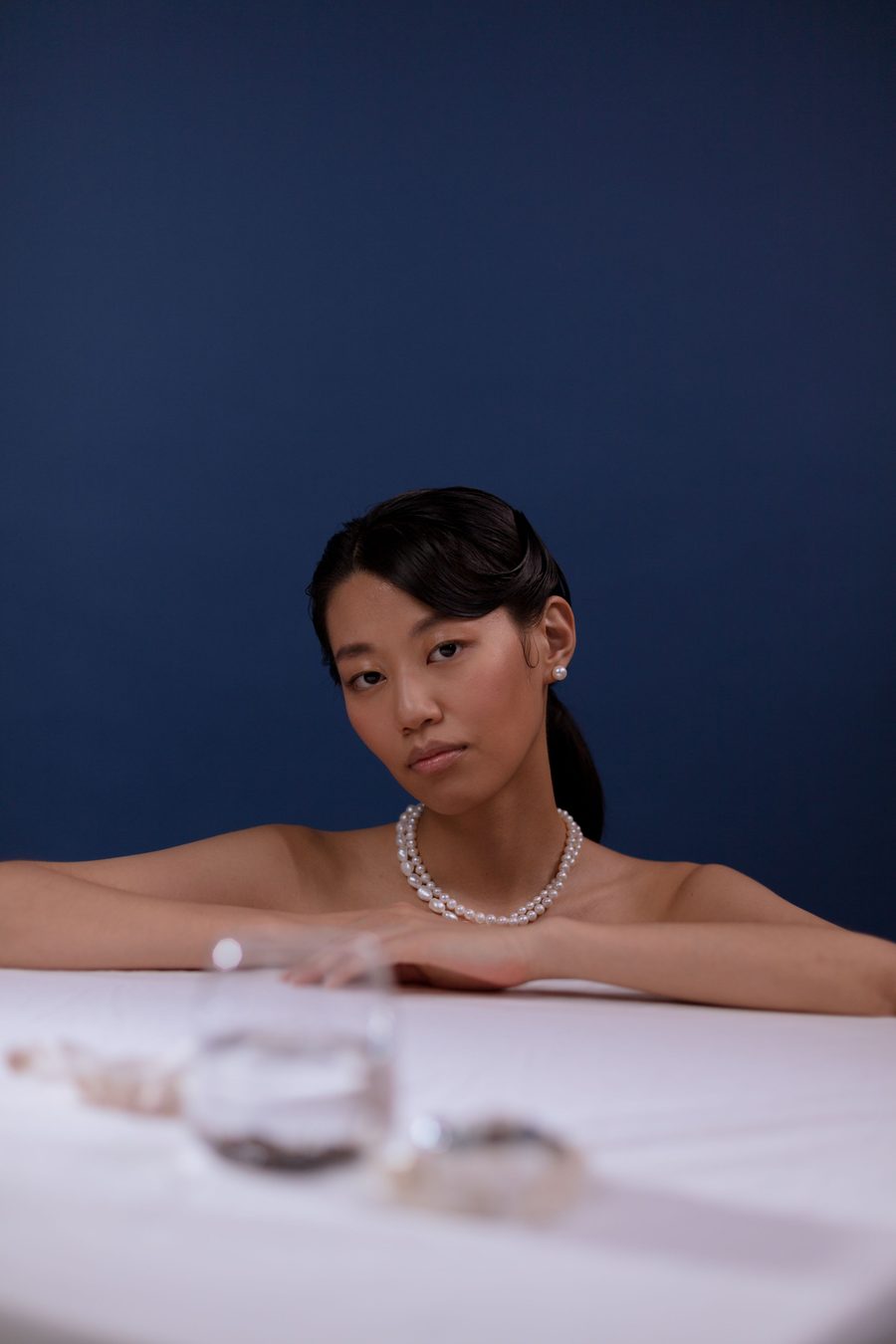 A woman wearing a pearl necklace and diamond stud earrings against a deep navy background.
